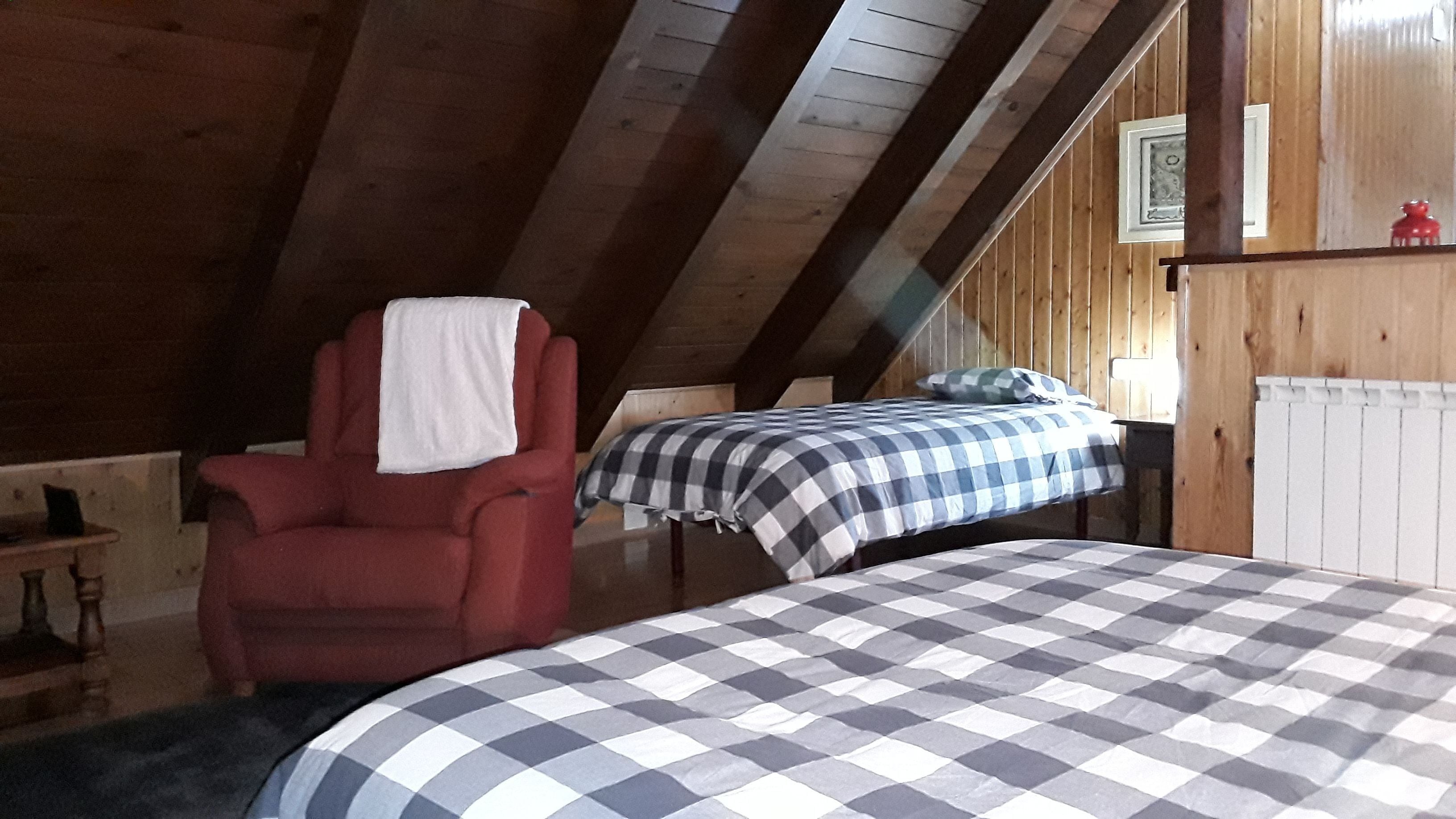 A room with a sloped ceiling, wooden walls, a red armchair with a white towel, a bed with a checkered bedspread, and a small table with a lamp.