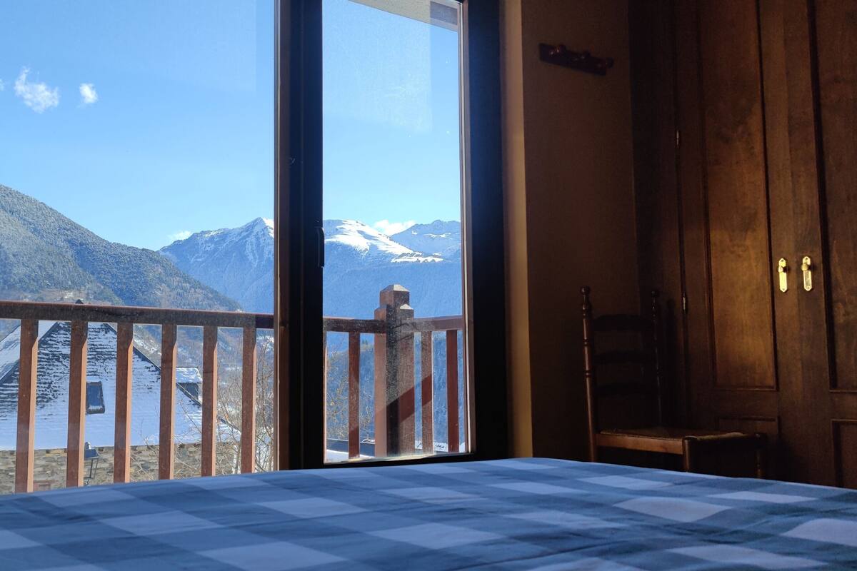 A room with a large window, a bed with a checkered bedspread, a wooden door, and a view of mountains.