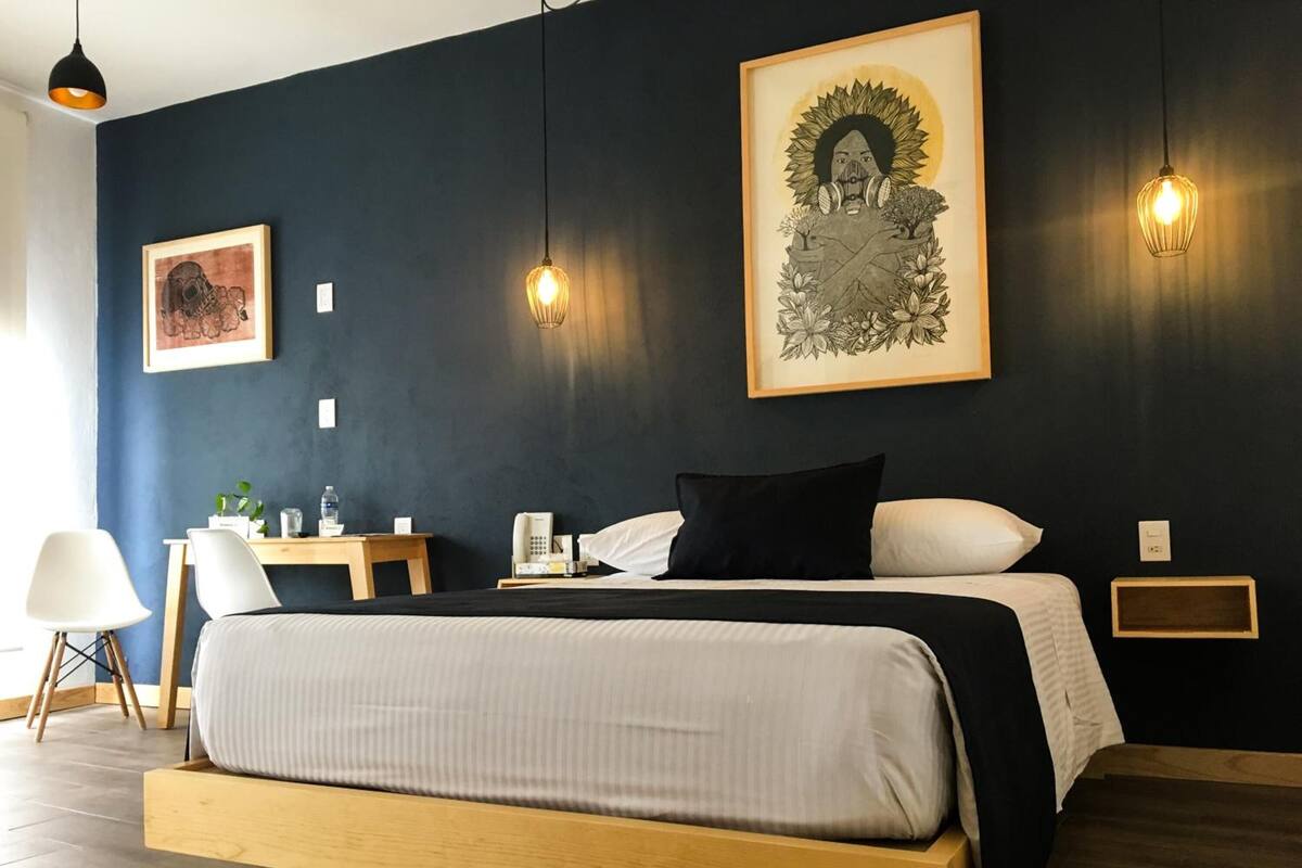 Una habitación de hotel moderna con una cama grande, mesitas de noche, un escritorio y una silla. Se observan obras de arte enmarcadas en las paredes y lámparas colgantes sobre la cama.