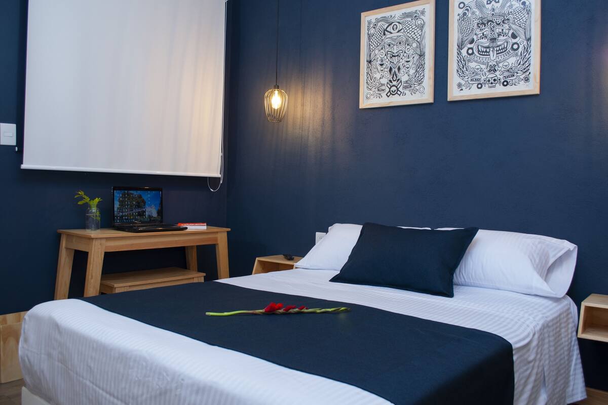Una cama bien hecha con una colcha azul oscuro y almohadas blancas, una mesita de noche con una lámpara, y dos obras de arte enmarcadas en la pared.