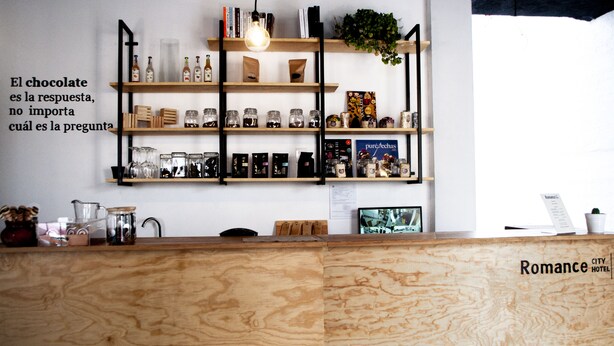 A reception desk with a wooden finish, shelves above displaying various items like books, bottles, and jars, and a sign with a quote in Spanish. - Romance City Hotel (Morelia)