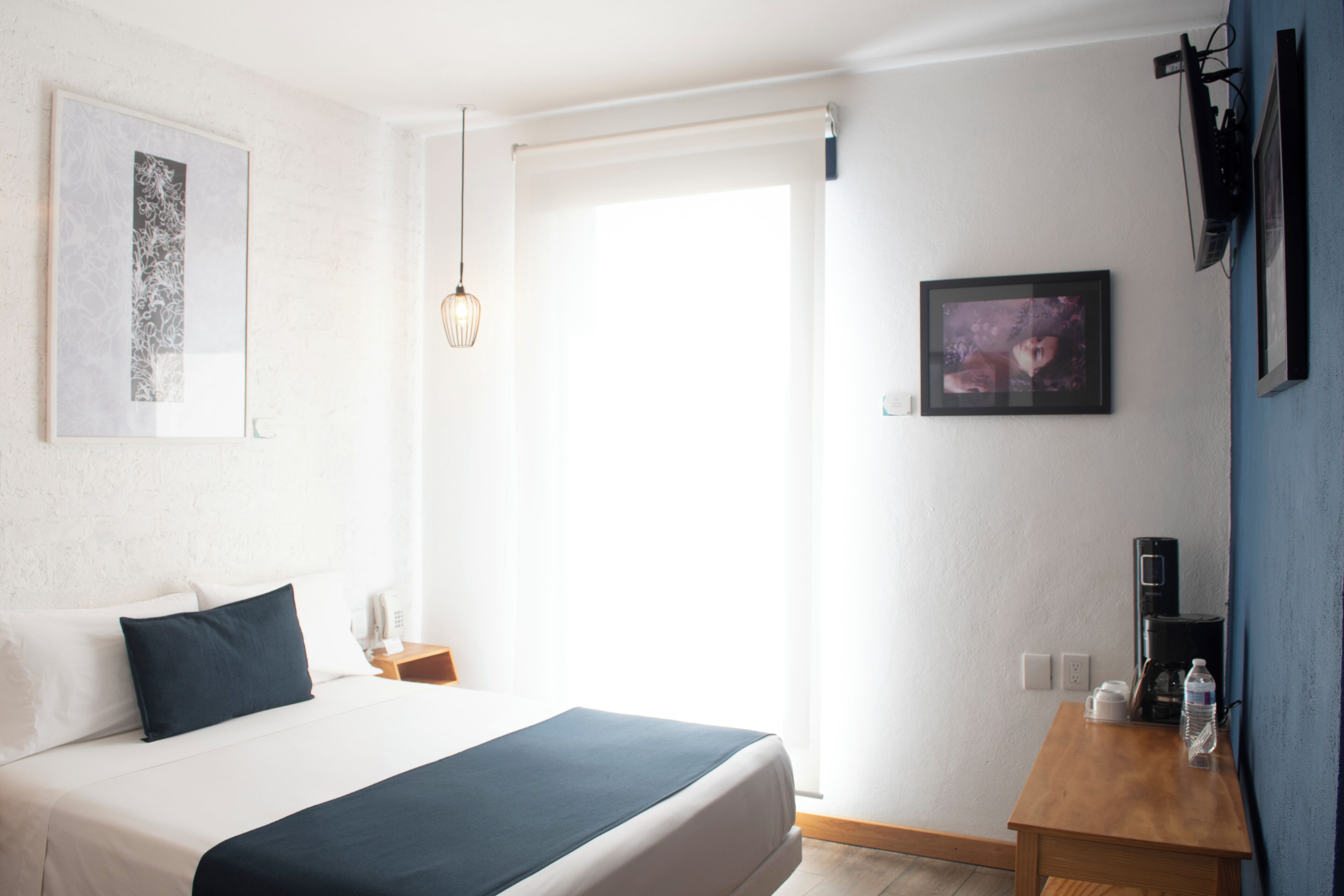 Ein Hotelzimmer mit einem Bett, einem Nachttisch mit Kaffeemaschine, einem Bild an der Wand und einem Fenster mit Vorhang.