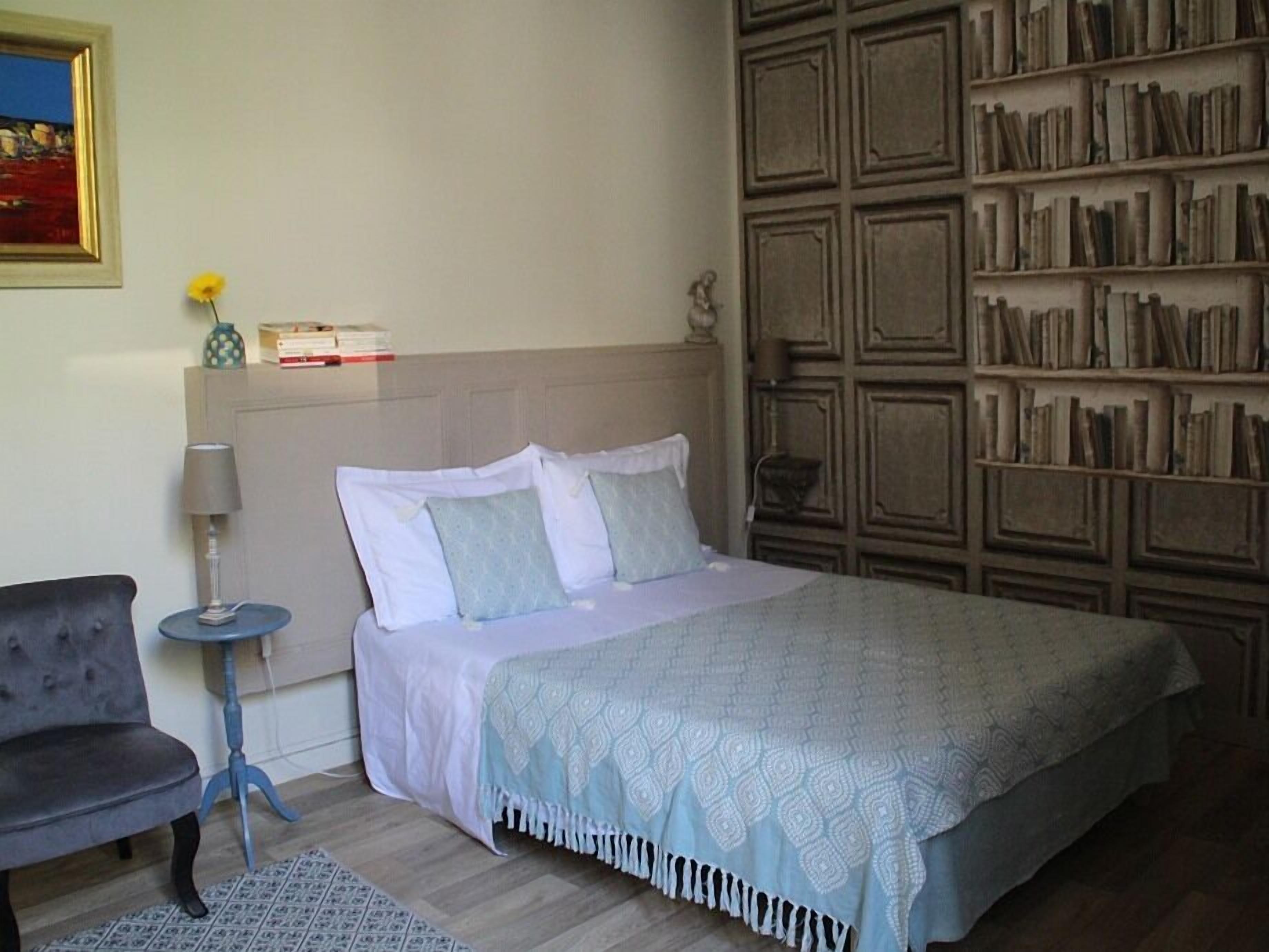 A bedroom with a bed, a chair, a small table, a bookshelf, and a painting on the wall.