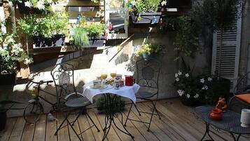 Een houten terras met metalen stoelen en een tafel gedekt voor een maaltijd, omgeven door potplanten en bloemen.