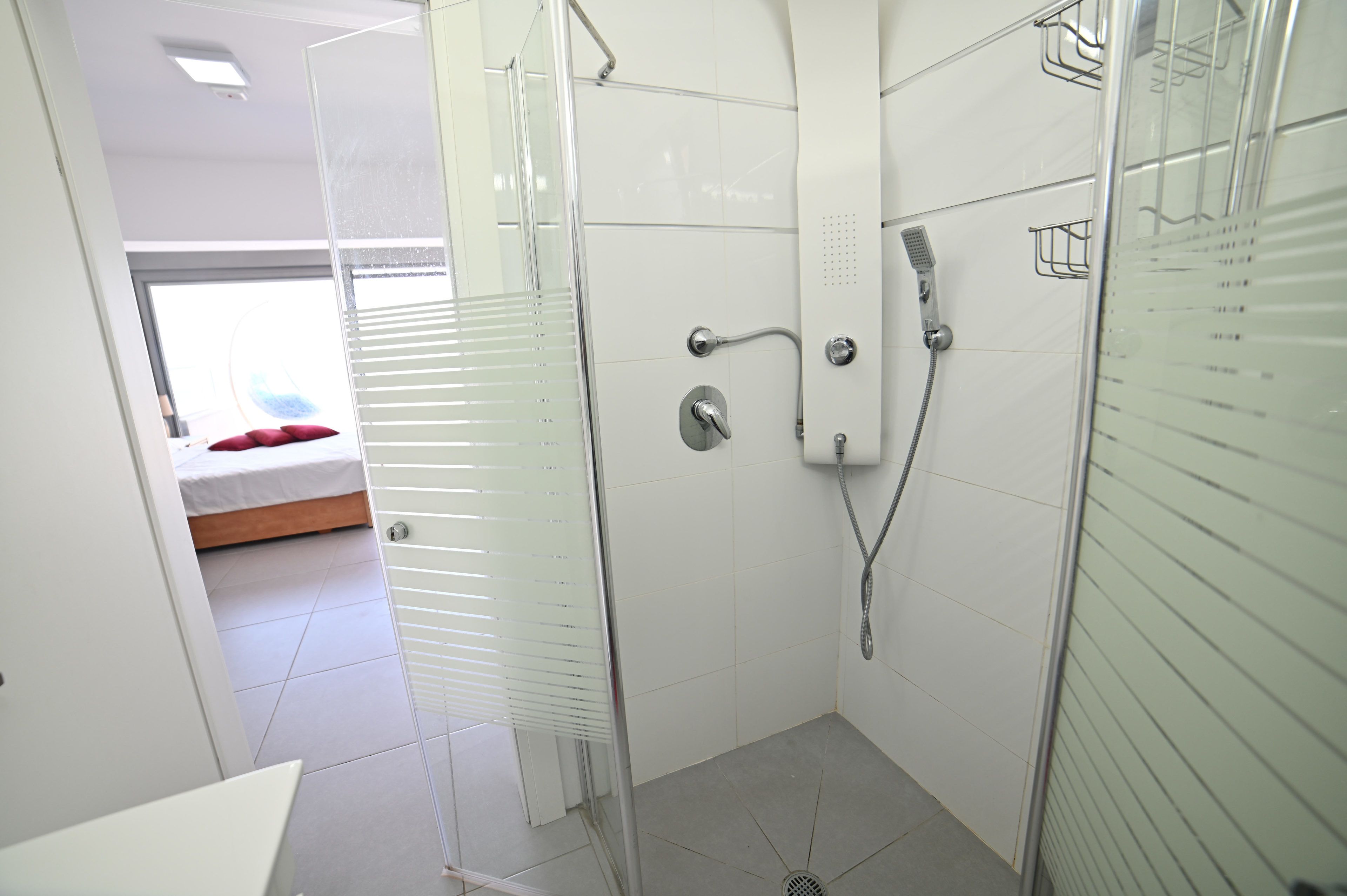 A modern bathroom with a glass shower enclosure, a visible toilet, and a bed in the adjacent room.