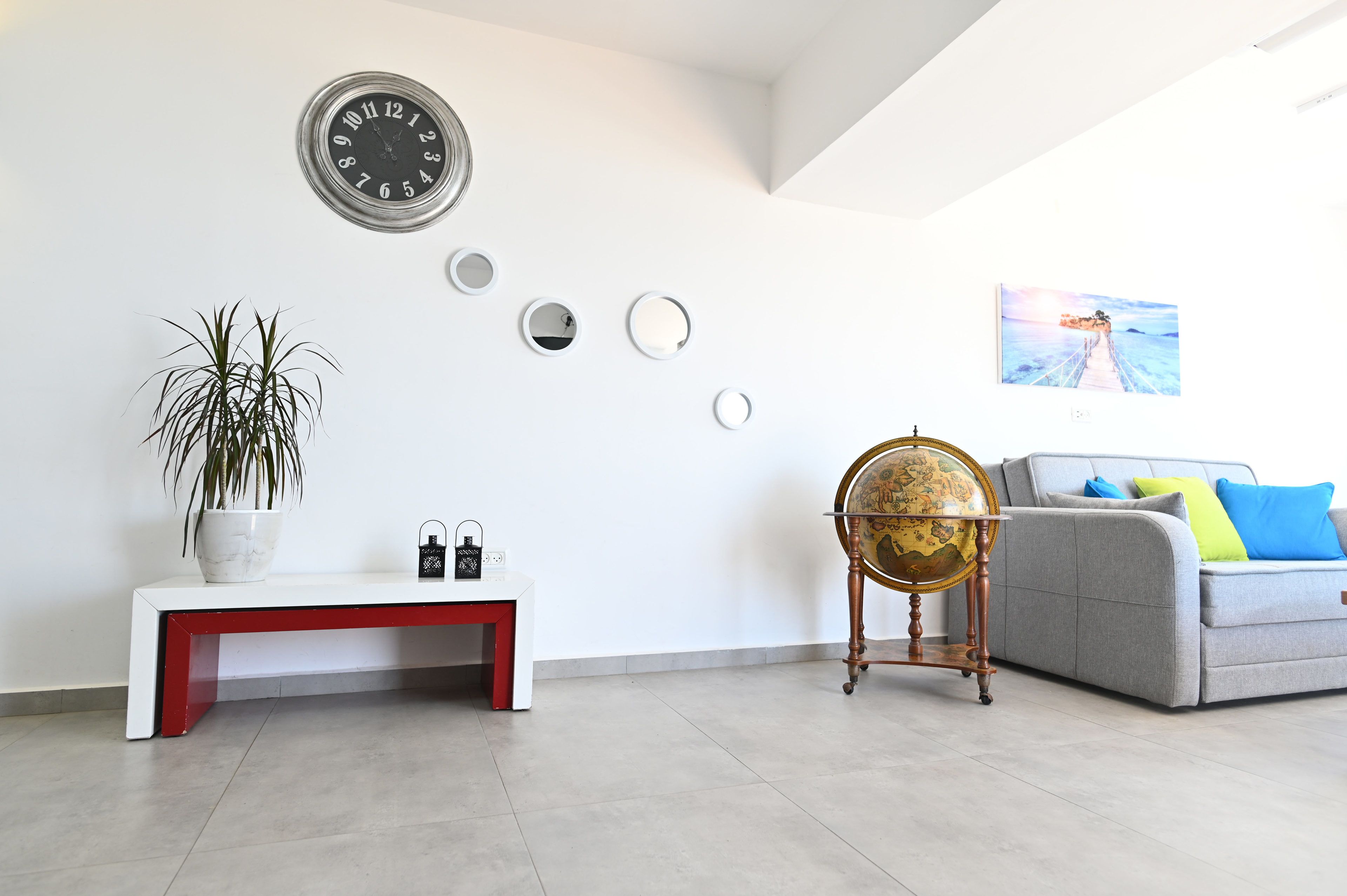 A modern living room with a white bench, a globe on a stand, a grey sofa with colorful cushions, a wall clock, and a framed painting.