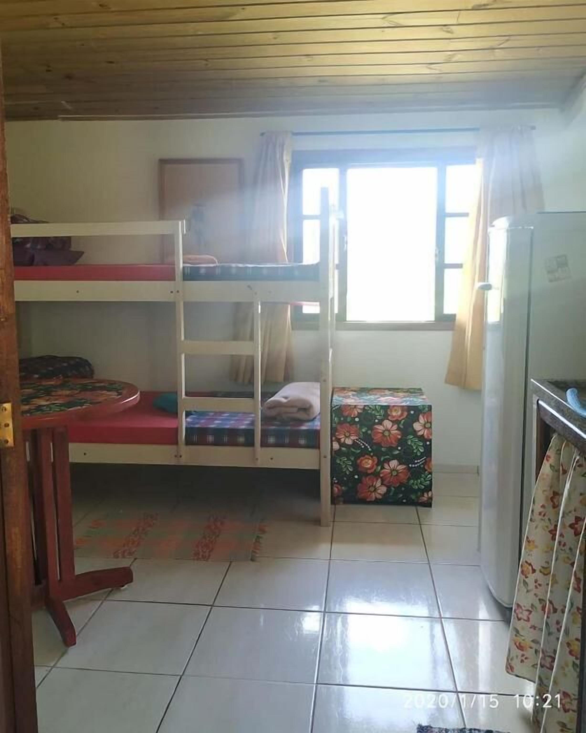 A dormitory room with bunk beds, a small table, a refrigerator, and a window with curtains.