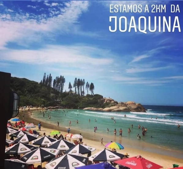 Uma praia com guarda-sóis e pessoas, com indicação da distância até Joaquina.