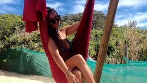 Una persona sentada en una hamaca roja sobre arena, con una red verde y vegetación al fondo.