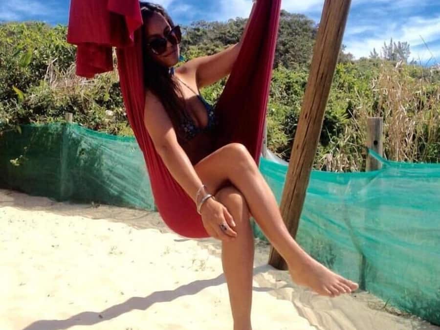 A person sitting in a red hammock on a sandy beach with green netting and vegetation in the background.