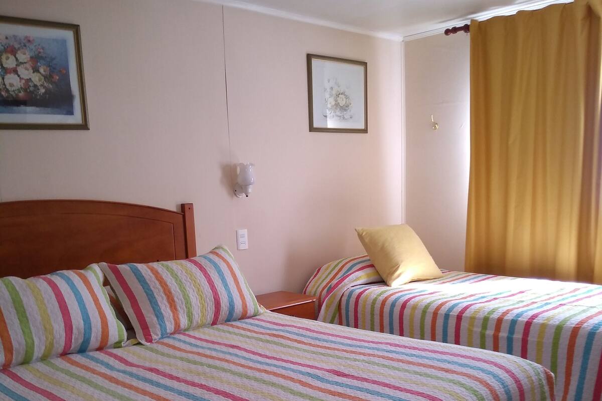 A hotel room with two beds, each with striped bedding, a wooden headboard, a nightstand, and a wall-mounted lamp.