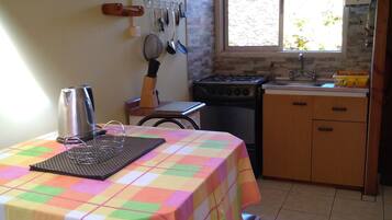 Una cocina con una mesa cubierta por un mantel colorido, una estufa, un fregadero y una ventana con vistas al verdor exterior.