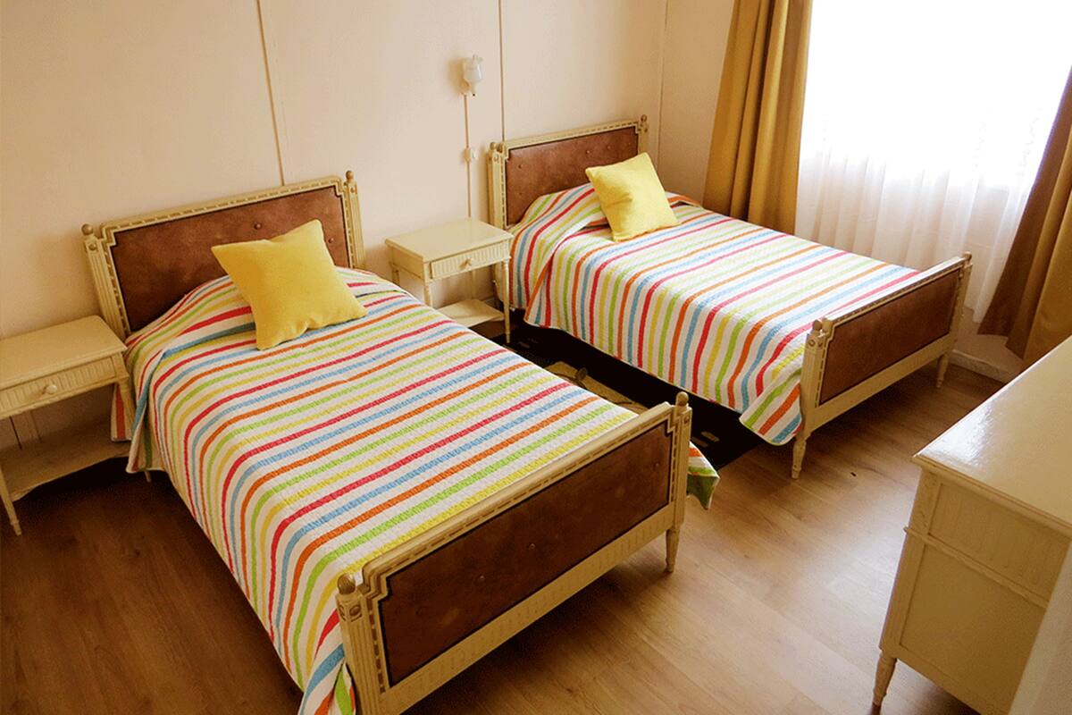 A room with two single beds, each with a wooden headboard and a colorful striped bedspread. There is a small bedside table with a lamp on each bed. The room has a wooden floor and a plain white wall with a light switch.