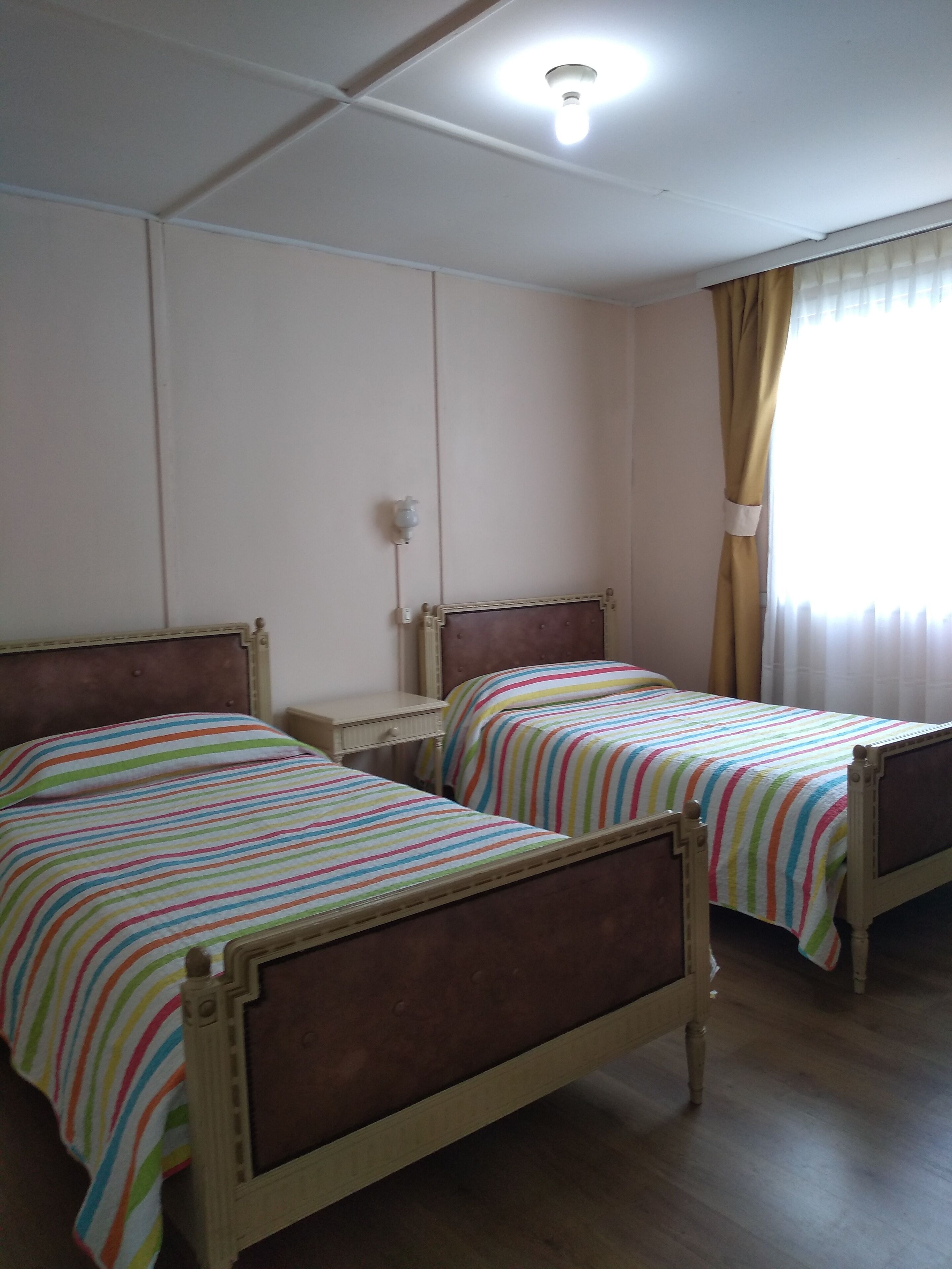 A room with two wooden beds with striped bedding, a bedside table, a wall-mounted light, and a window with curtains.