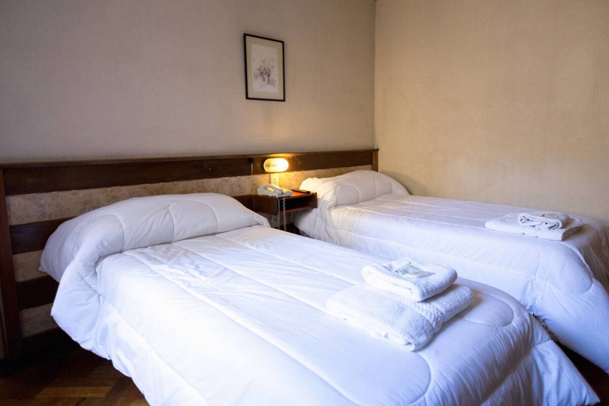 A hotel room with two beds, a bedside table with a lamp, and a framed picture on the wall.