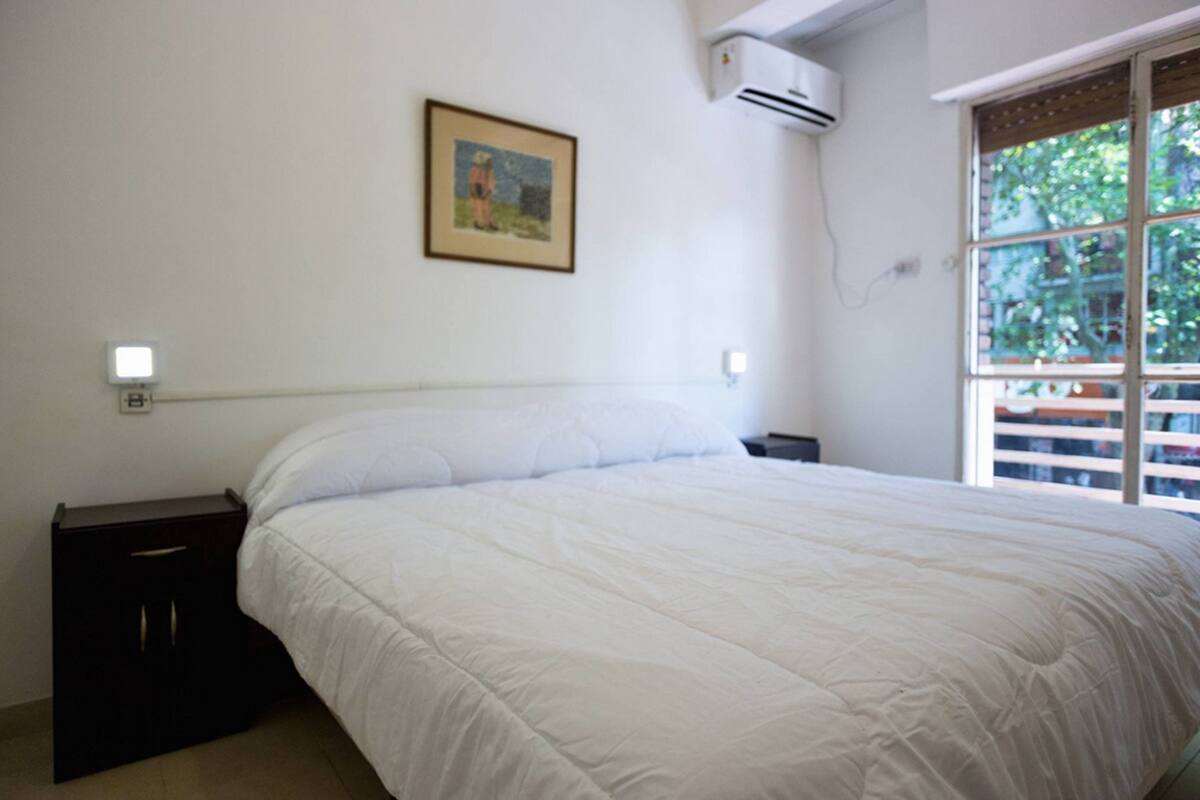 A bedroom with a large bed, a nightstand, a wall-mounted air conditioner, a window with a view of greenery, and a framed picture on the wall.