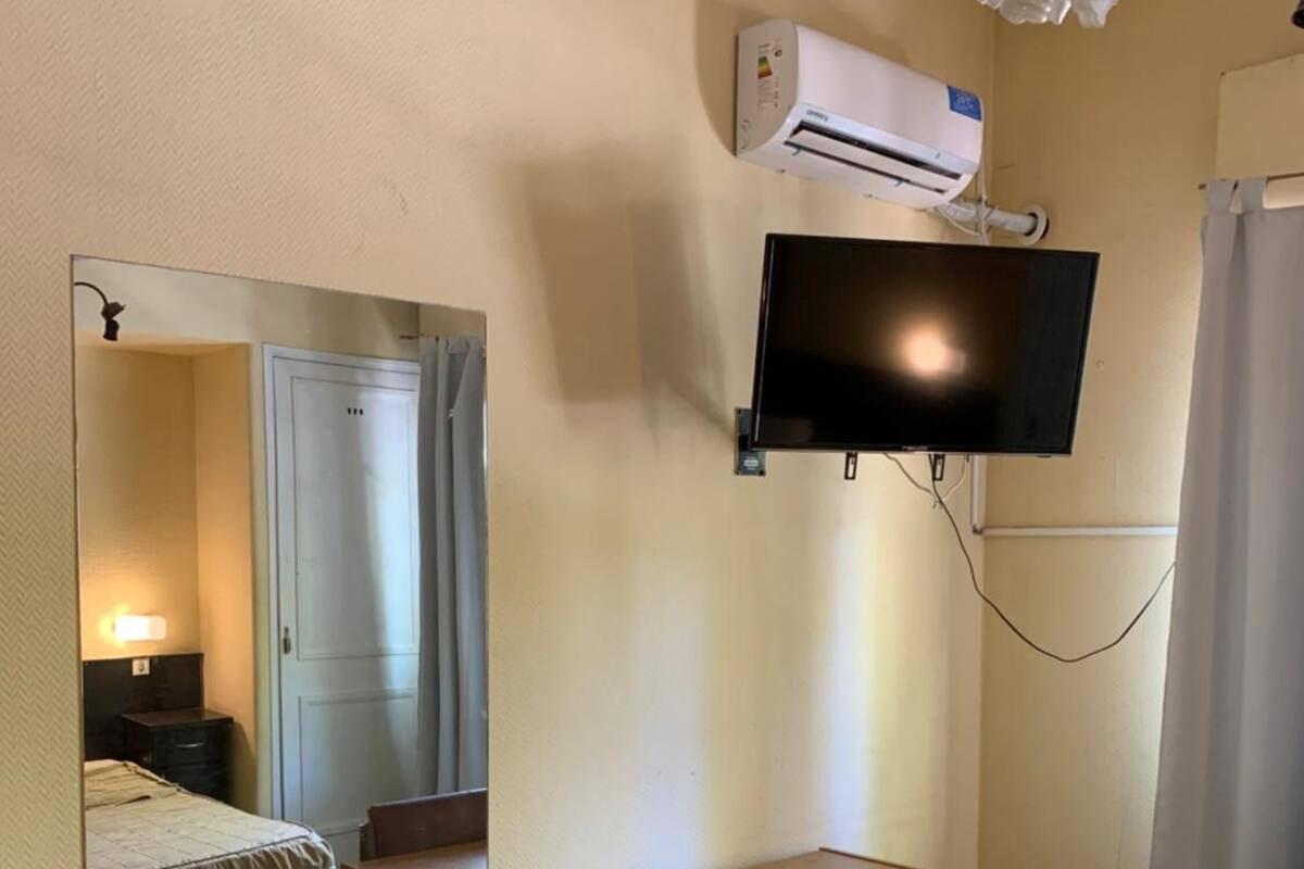 A hotel room with a bed, a desk with a chair, a flat-screen TV mounted on the wall, an air conditioning unit, and a mirror reflecting the room.