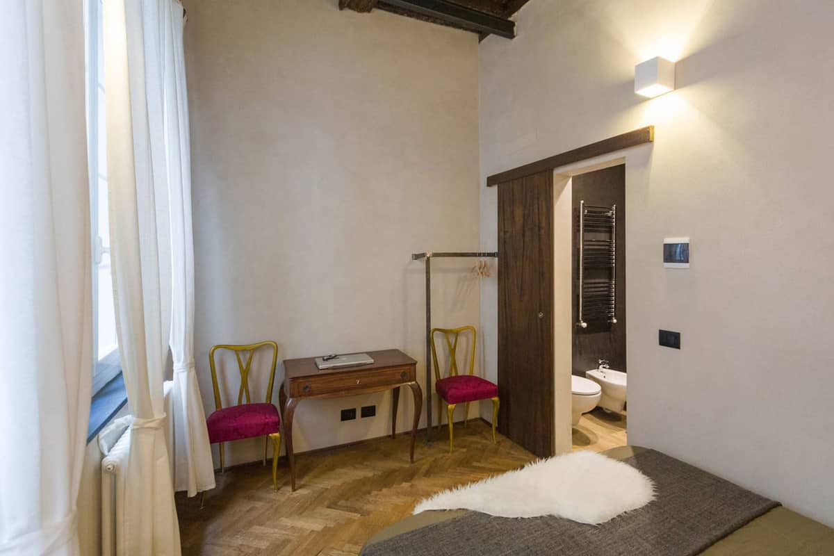 A small, cozy room with a wooden floor, a desk with two chairs, a white fur rug, and a bathroom with a toilet and bidet.