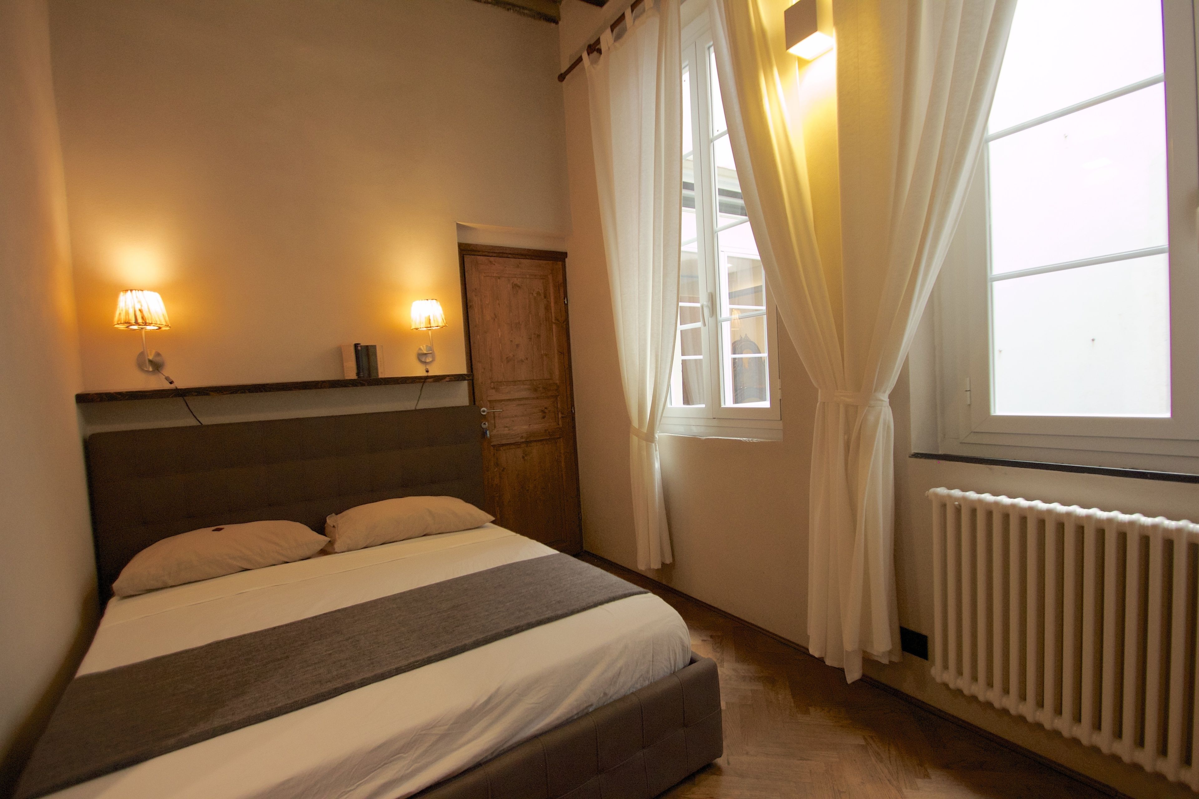 Ein Schlafzimmer mit einem Bett, zwei Wandleuchten, einem Holzschrank, einem Fenster mit Vorhängen und einer Heizung.