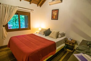 Un dormitorio con cama, mesita de noche, ventana con cortinas, un cuadro en la pared y suelo de madera.