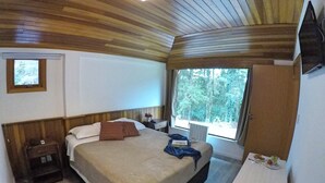 A bedroom with a wooden ceiling, a bed with a beige bedspread, a nightstand, a window with a view of trees, and a small table with a cup and a plate. - Pousada Rosa Gramado (Gramado)