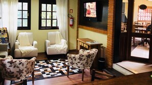 A cozy living room with two white armchairs, a patterned armchair, a small wooden table, and a television on the wall. - Pousada Rosa Gramado (Gramado)