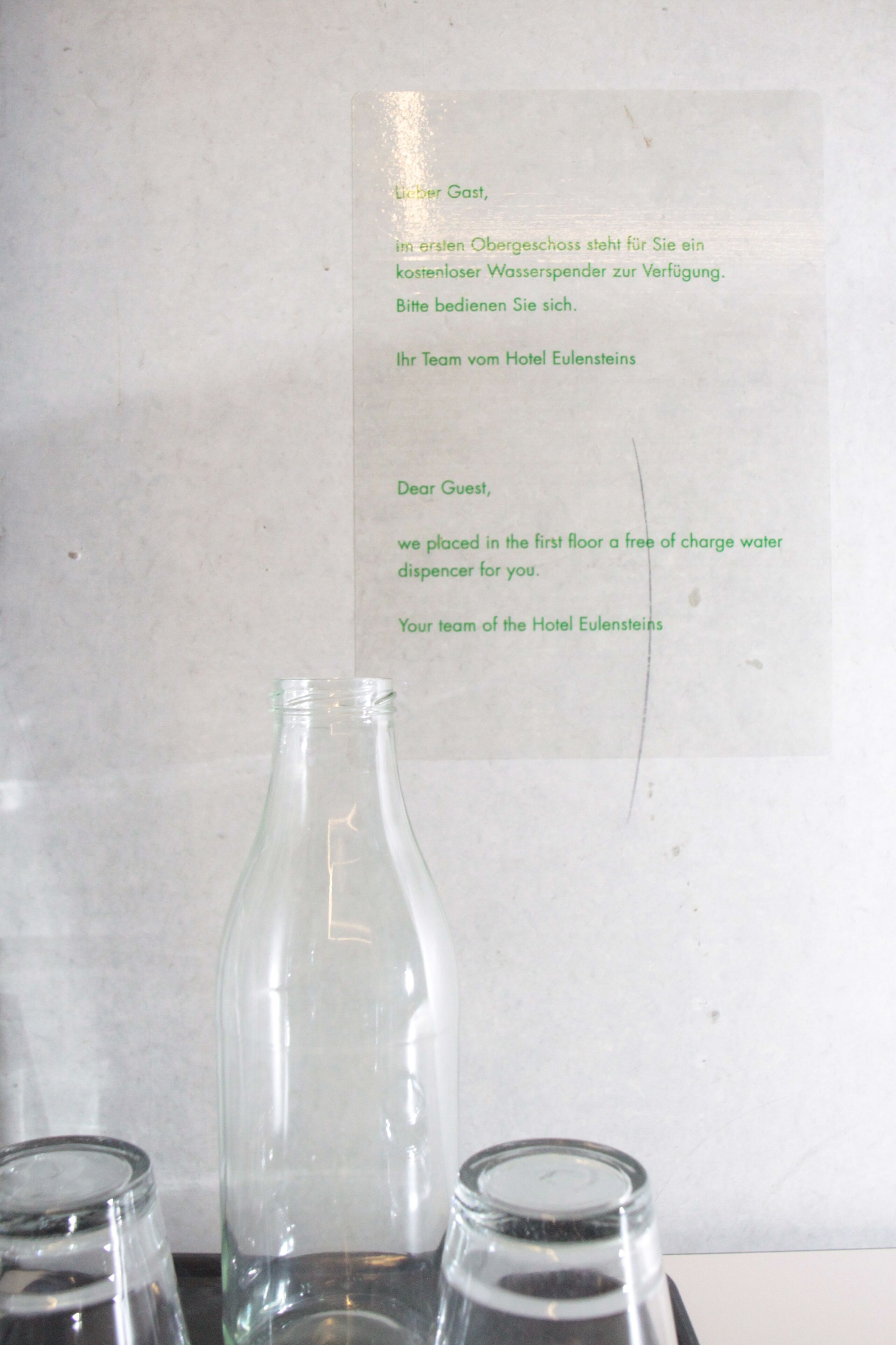 A hotel sign in German and English, a clear glass bottle, and two empty glasses on a table.
