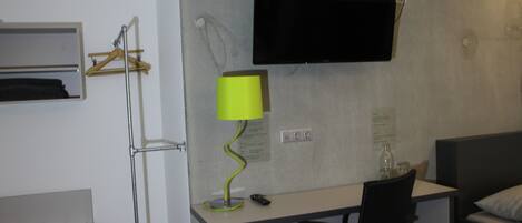 A hotel room with a desk, a chair, a bed, a TV mounted on the wall, a lamp, and a coat rack.