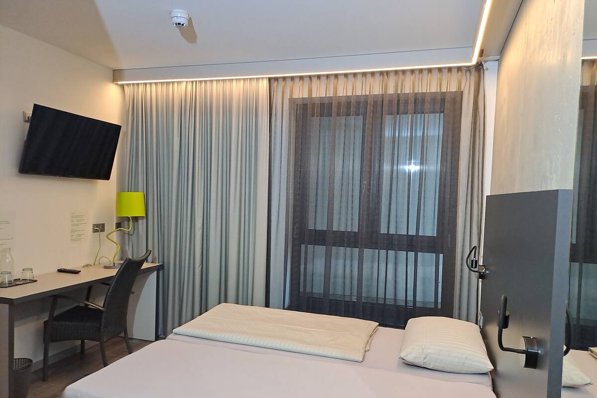 A hotel room with a bed, a desk, a chair, a TV mounted on the wall, and a window with curtains.