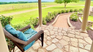 A porch with a wicker chair, a stone pathway, and a view of a green field and tree. - Southern Backroad Retreats (The Pool House) (Goshen)