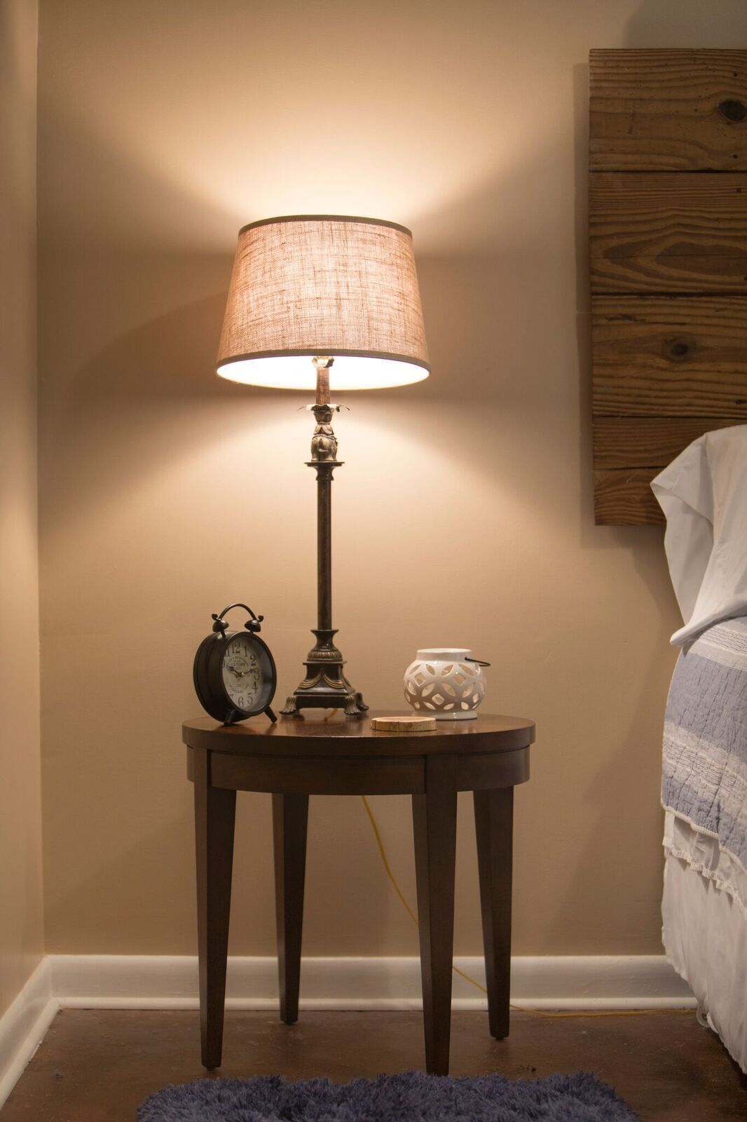 A bedside table with a lamp, an alarm clock, and a decorative item.