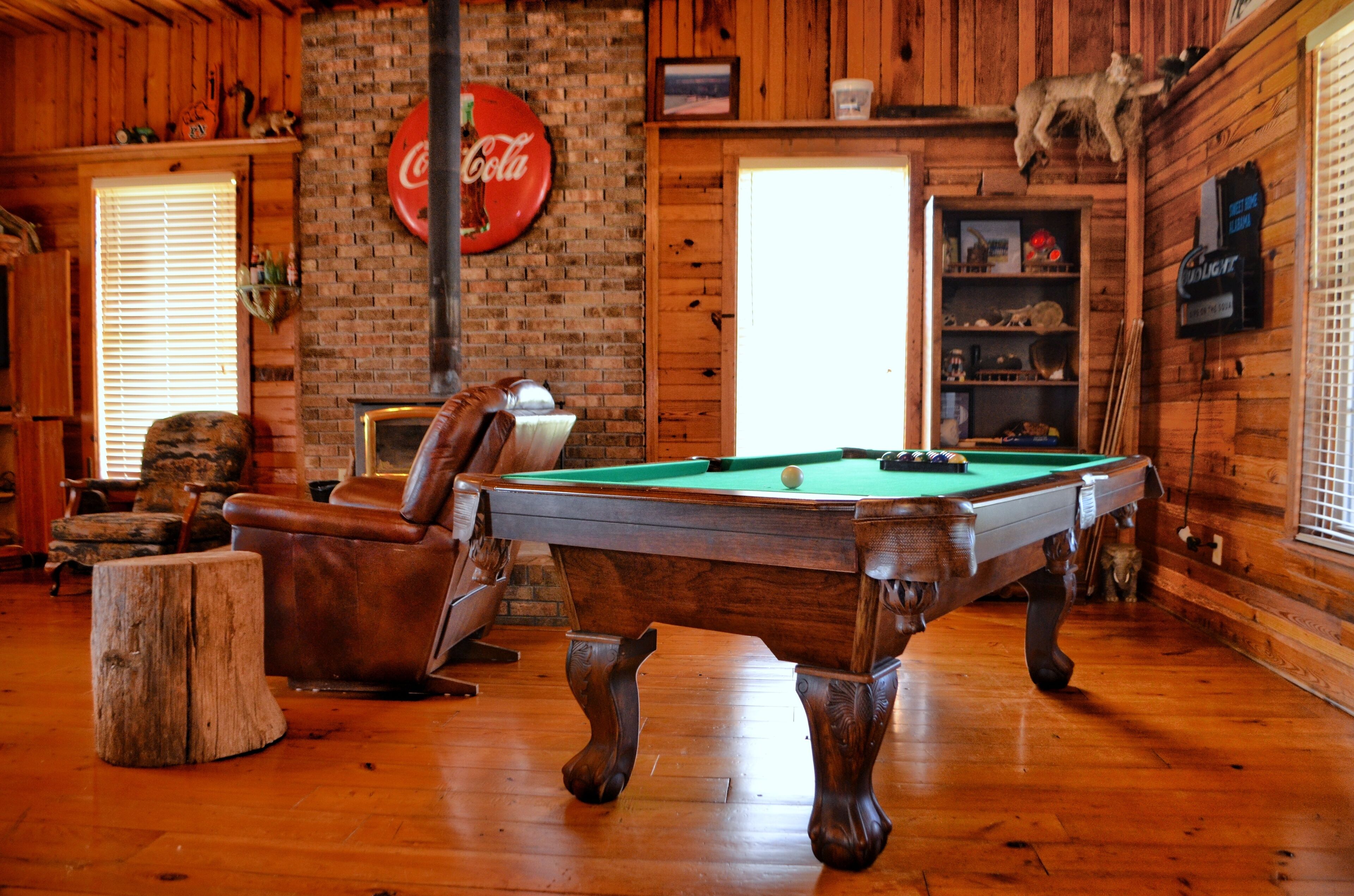 A wooden interior with a pool table, leather chairs, a brick wall, and a Coca-Cola sign.