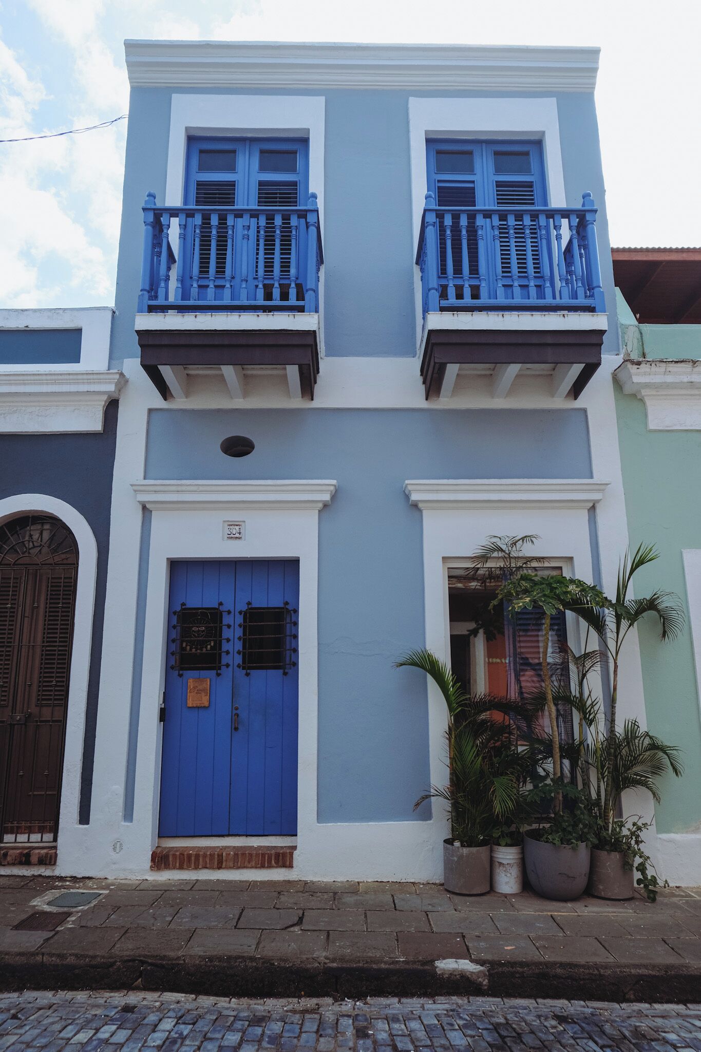 Un immeuble de deux étages avec une porte bleue et un balcon, des plantes en pot au rez-de-chaussée et le numéro « 304 » sur la porte.