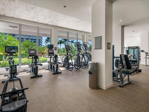 A modern gym with cardio equipment, weight machines, and a view of palm trees and a building.