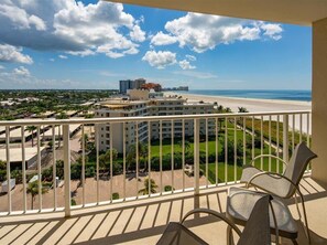 A balcony with wicker chairs overlooking a beach and a hotel building.