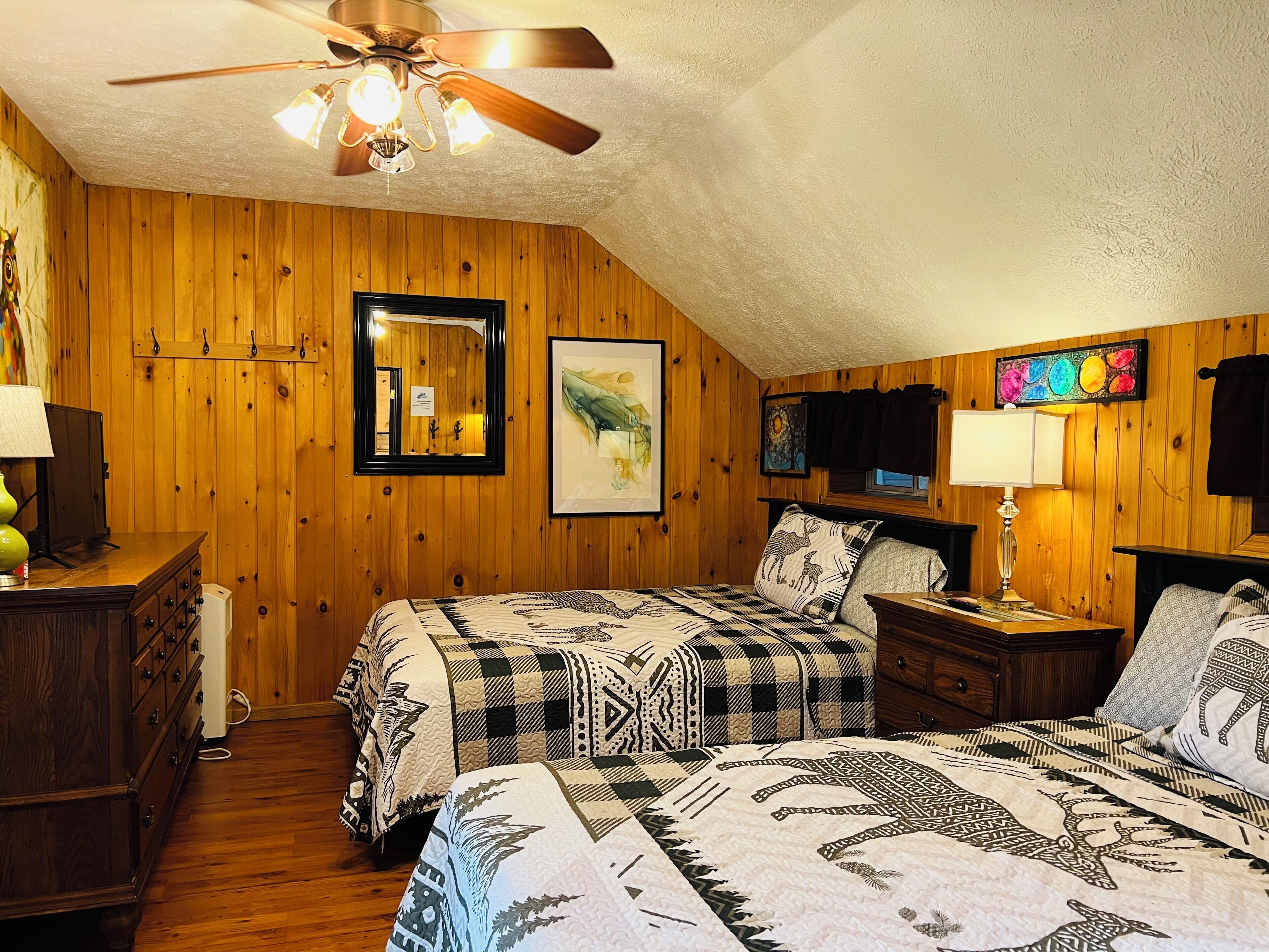 Een kamer met houten wanden, een plafondventilator, een bed met beddengoed met patroon, een dressoir, een nachtkastje en een ingelijst beeld aan de muur.