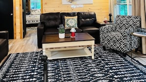 Interior de una cabaña de madera con ventilador de techo, un sofá de cuero negro, una mesa de centro, un tapete estampado y una ventana con cortinas.