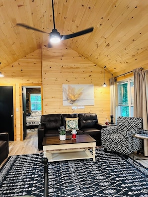 A wooden cabin interior with a ceiling fan, a black leather sofa, a coffee table, a patterned rug, and a window with curtains.