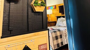 Una habitación con una puerta de madera oscura, un letrero de bienvenida, macetas hexagonales de madera con suculentas, una cama con colcha a cuadros y un cartel que indica un albergue.