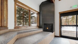 A modern interior with a fireplace, wooden steps, and large windows offering a view of trees.