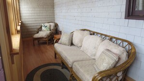 Una habitación con techo de madera, pared de ladrillo blanco, sofá de mimbre, una silla de madera y una pequeña mesa de madera.