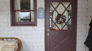 Una puerta con un adorno festivo, una ventana con cortina y una silla de mimbre.
