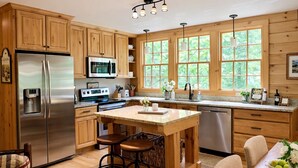 A kitchen with wooden cabinets, stainless steel appliances, a central island with stools, and large windows with a view of greenery.