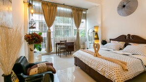 Une chambre d’hôtel avec un lit, un bureau et une chaise. Il y a une fenêtre avec des rideaux, une lampe et un vase rempli de fleurs.