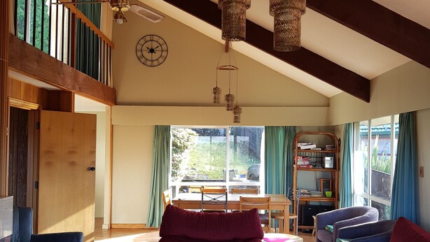 A spacious living room with a high ceiling, wooden beams, and a large window. There are two red sofas, a blue armchair, and a wooden coffee table. A dining area with a table and chairs is visible in the background. - Cosy spacious Chalet Experience. Beautiful views. (Hanmer Springs)