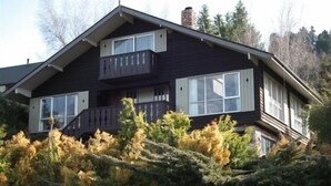 A two-story house with a balcony on the upper floor, surrounded by dense vegetation. - Cosy spacious Chalet Experience. Beautiful views. (Hanmer Springs)