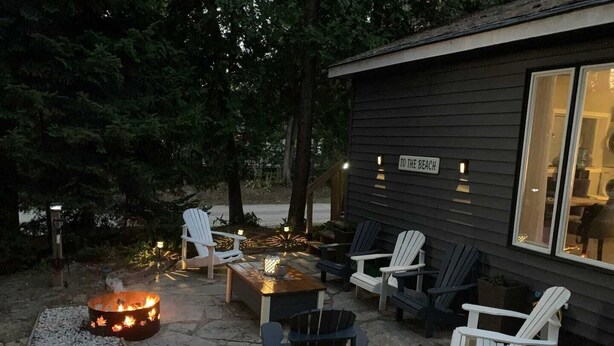 Cómoda zona de estar al aire libre con sillas Adirondack y un brasero, rodeada por una terraza de madera y una casa con un letrero que dice 'TO THE BEACH'.