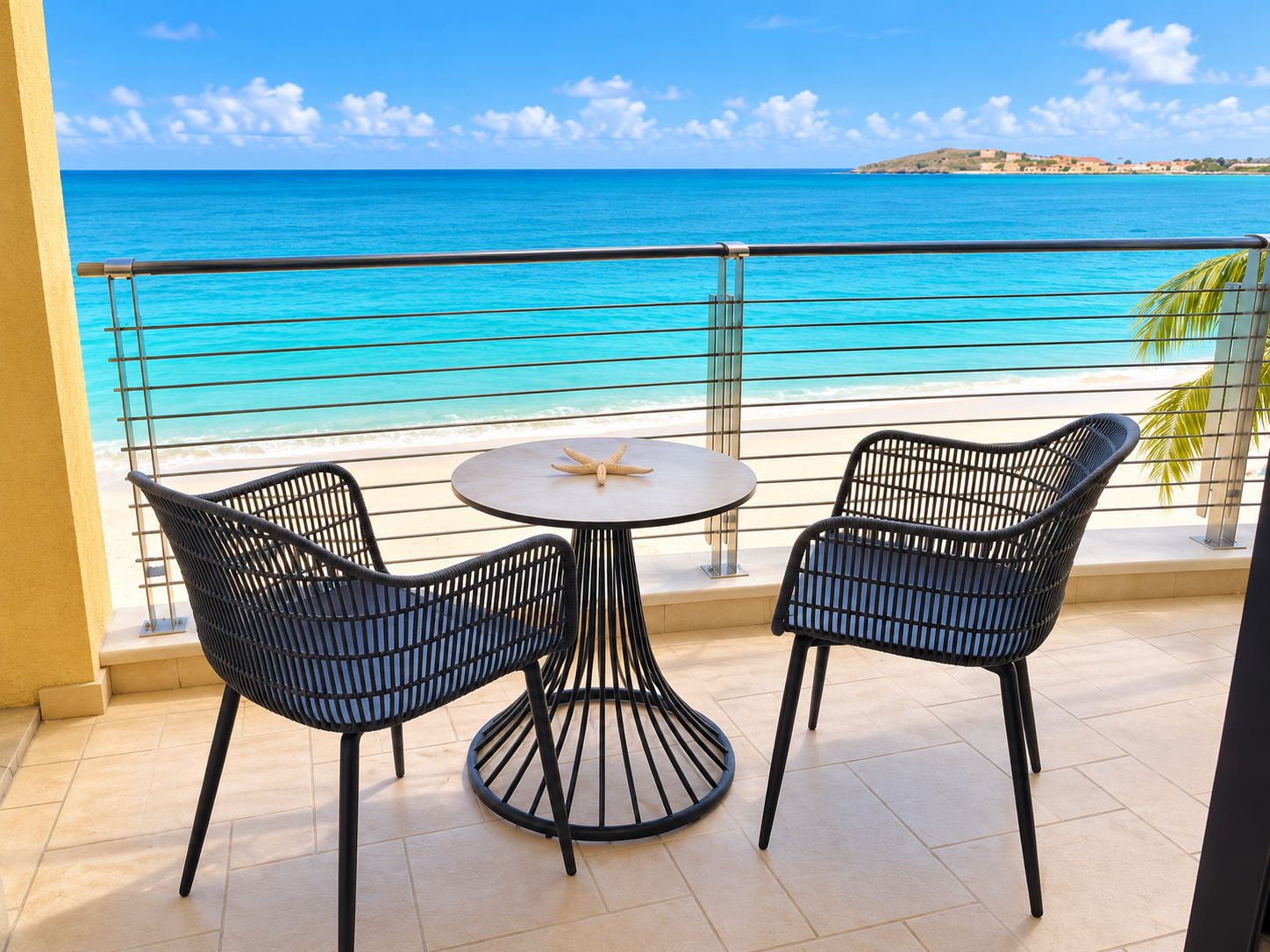 Un balcon avec deux fauteuils en osier et une petite table ronde, donnant sur une plage aux eaux d’un bleu limpide et sur une île lointaine.