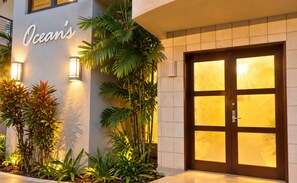 A hotel entrance with a large wooden door, a sign reading 'Ocean's', and well-maintained tropical plants.