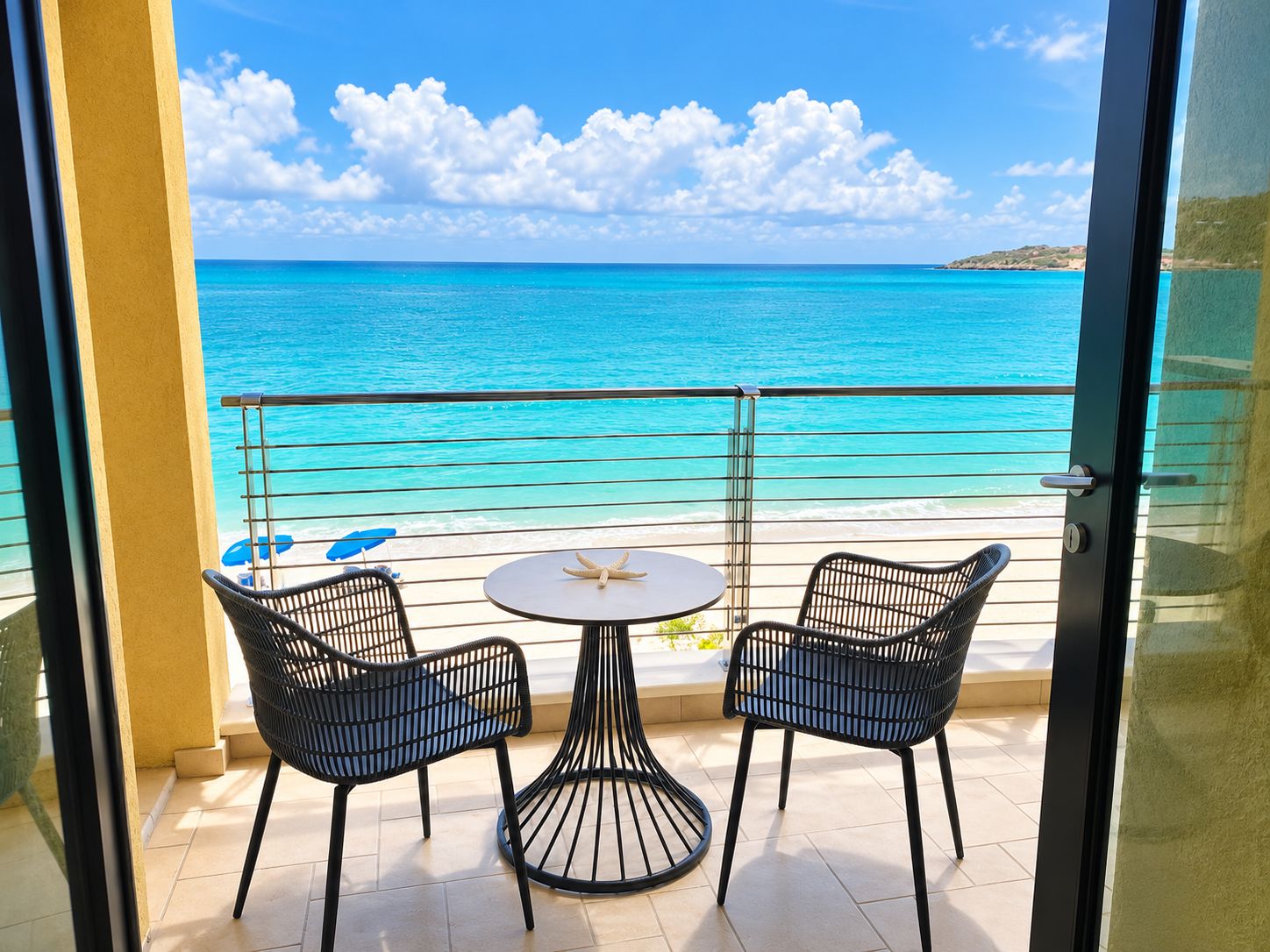 A balcony with two chairs and a table, overlooking a beach with clear turquoise water and a distant island.